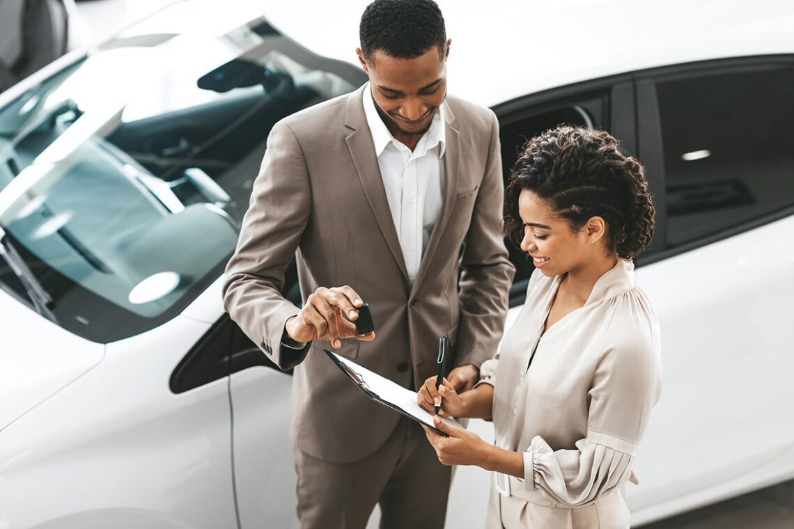 Dealer and customer with paperwork, a keyfob, and a white car
