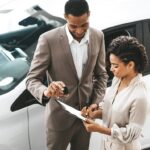 Dealer and customer with paperwork, a keyfob, and a white car